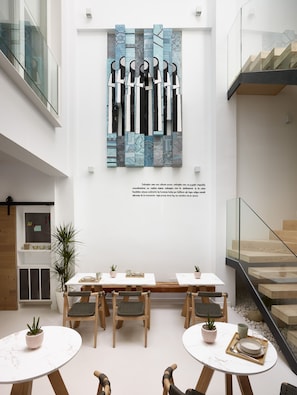 A modern interior with a minimalist design, featuring a dining area with wooden tables and chairs, potted plants, and a large abstract art piece on the wall.