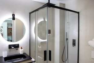 Un baño moderno con ducha envuelta en vidrio, un espejo redondo y un lavabo negro.