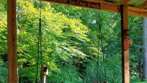 Un ensemble de balançoires en bois installé sous une pergola, agrémenté d’une pancarte indiquant « Bienvenue sur notre véranda », le tout entouré d’une végétation luxuriante.