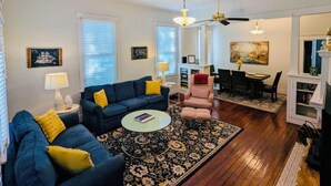 A living room with a blue sofa, a pink armchair, a glass coffee table, and a dining area with a table and chairs. - Magnolia Beach House l Brand NEW AC UNIT (Gulfport)