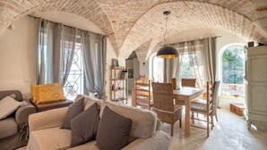 Ein gemütliches Wohnzimmer mit Backstein-Decke, einer Couch, einem Esstisch mit Stühlen und Blick durch die Fenster.