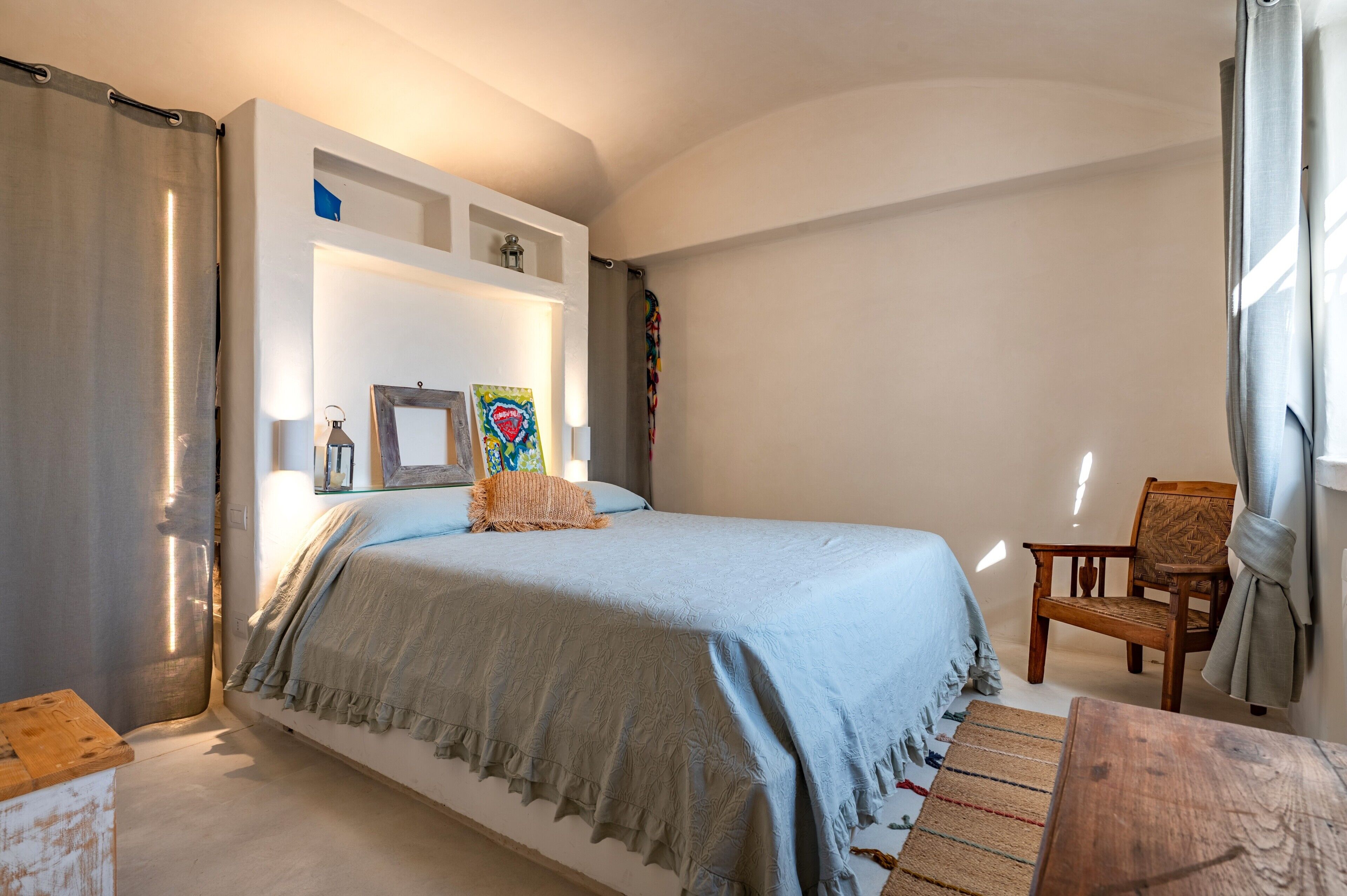 A bedroom with a bed, a chair, a small table, and a shelf with decorative items.