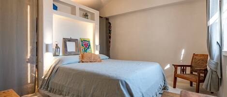 A bedroom with a bed, a chair, a small table, and a shelf with decorative items.