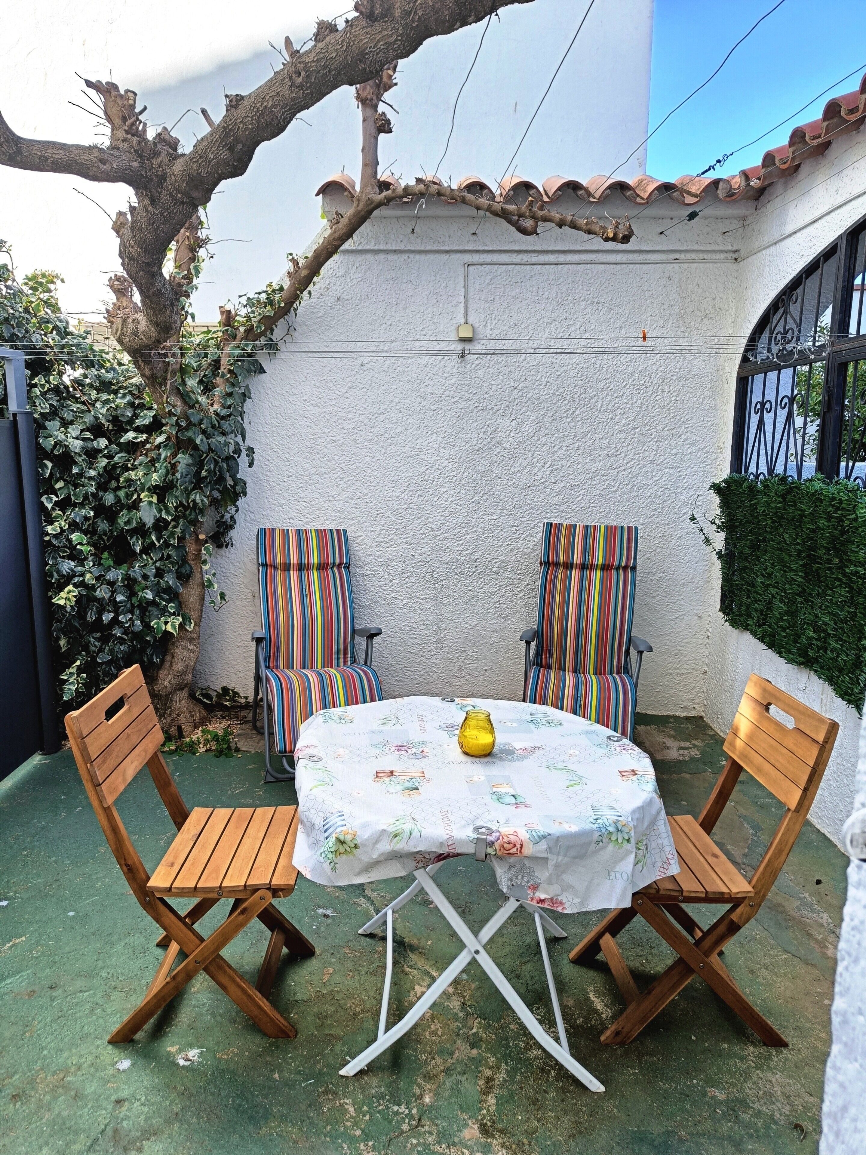 Une terrasse avec une table et deux chaises à rayures, un vase jaune et un arbre recouvert de lierre.