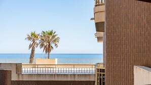A balcony with a view of the sea, palm trees, and a brick building.