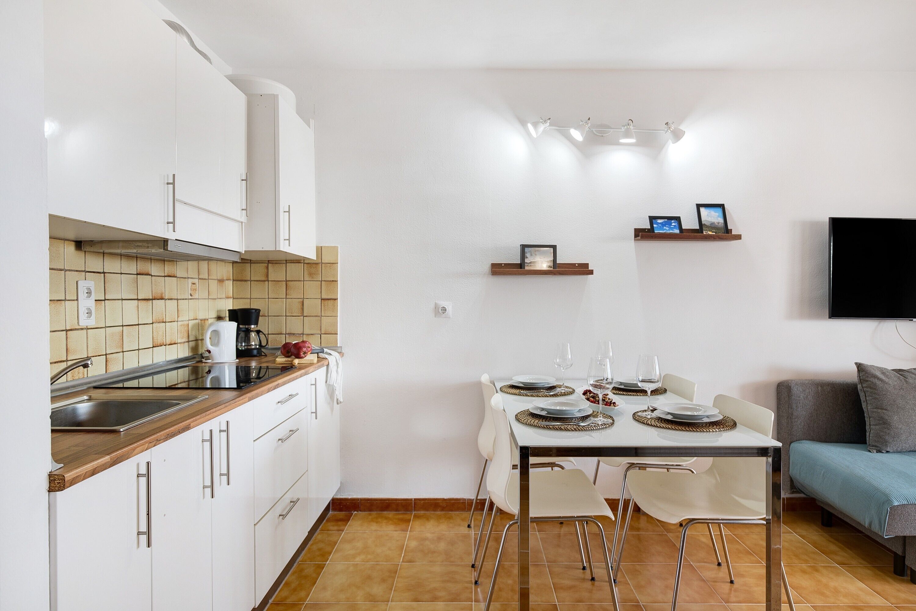 A compact kitchen with white cabinets, a dining table set for four, and a flat-screen TV mounted on the wall.