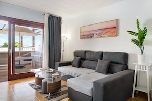 A modern living room with a grey sofa, a glass coffee table, a white side table with a plant, and a painting of a sunset on the wall.