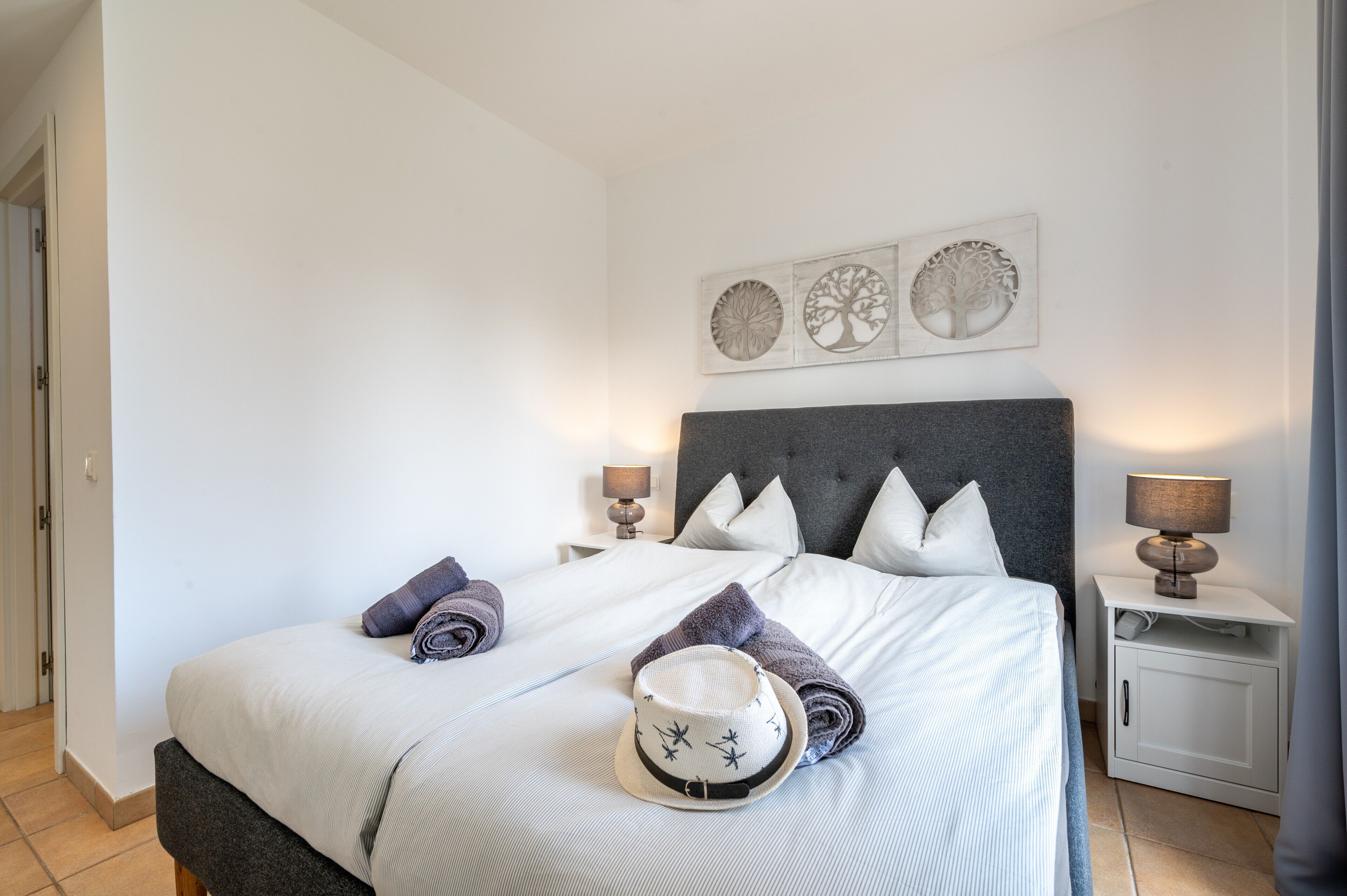 Un lit bien fait, avec des oreillers, un chapeau et des serviettes. Deux œuvres d’art sont accrochées au mur au-dessus du lit.