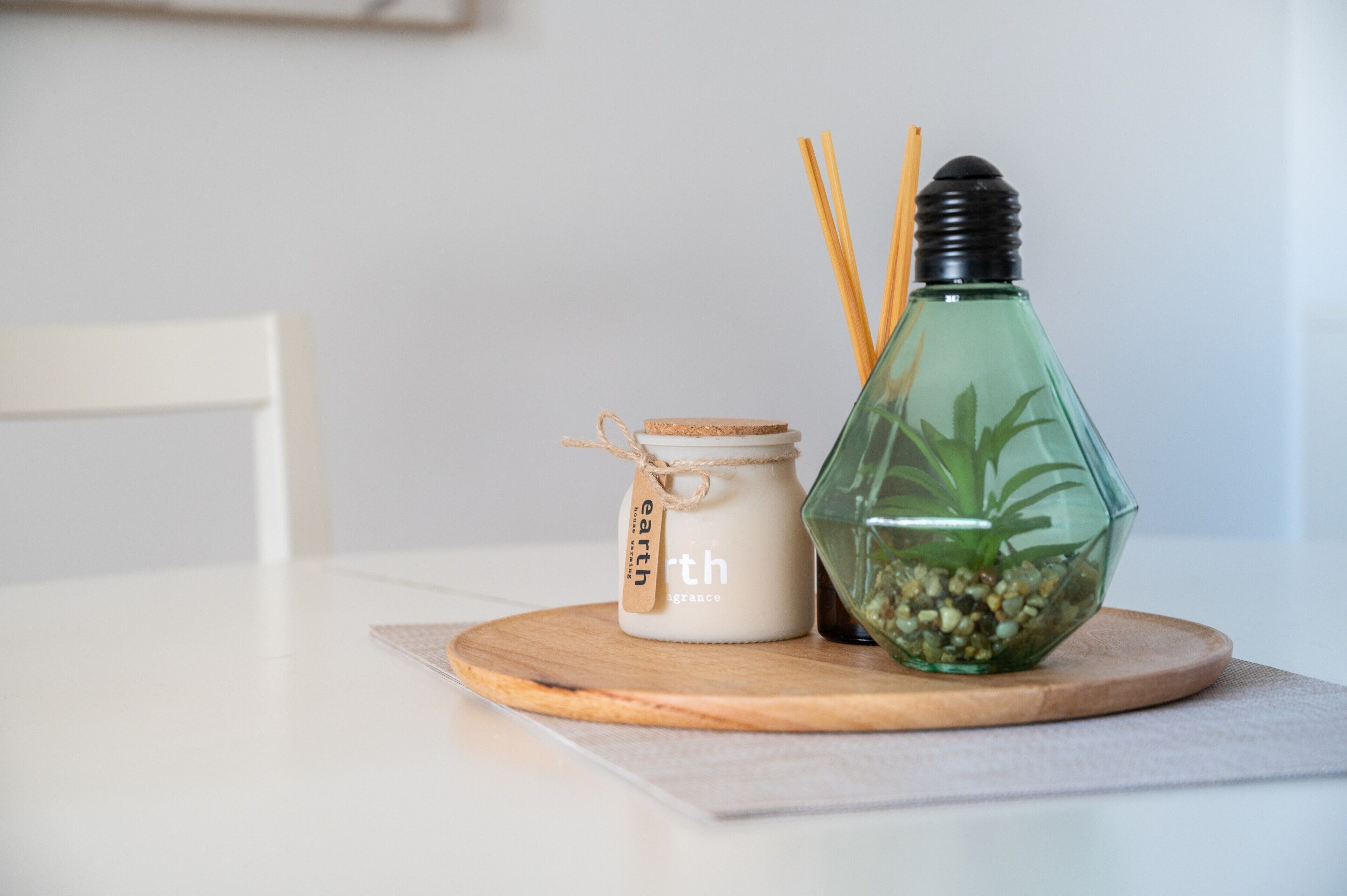Et lys i et glas, en mærkeetiket, en glasflaske med en plante og to træpinde på en rund træunderlag.