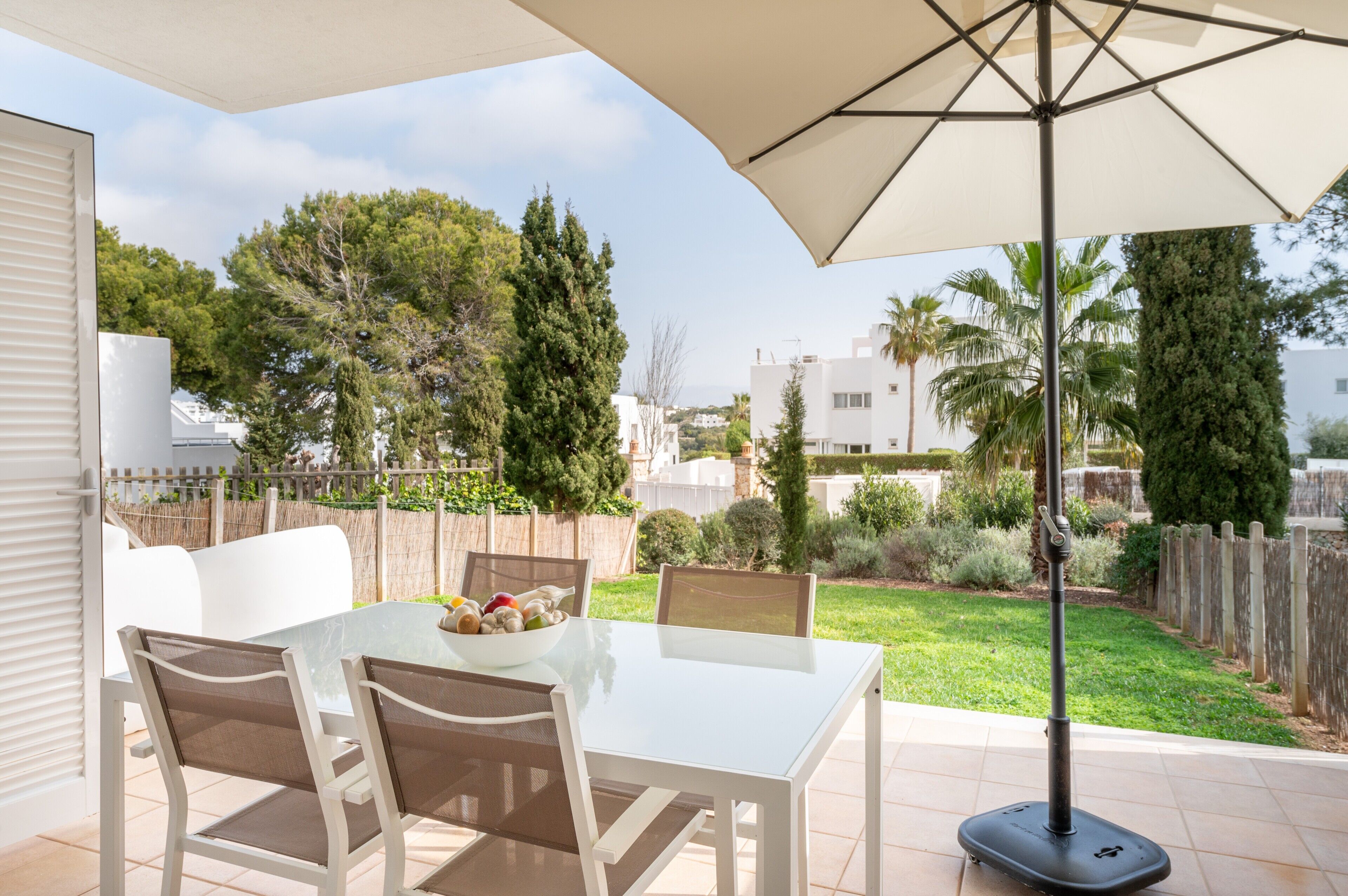 Eine Terrasse im Freien mit Tisch und Stühlen, einem großen Sonnenschirm und Blick auf einen Garten sowie Gebäude im Hintergrund.