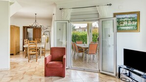 A spacious living area with a dining table, chairs, a red armchair, a TV, and a glass door leading to an outdoor patio. - Villa 'Casa Flores' with Mountain Views, Shared Pool, and Wi-Fi (Vilacolum)