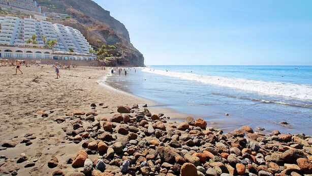 A beach with rocky foreground, a white building with balconies, and a clear blue sky. - Vacation Apartment 'Paraíso Taurito' with Sea View, Terrace, Shared Pool & Wi-Fi (Taurito)