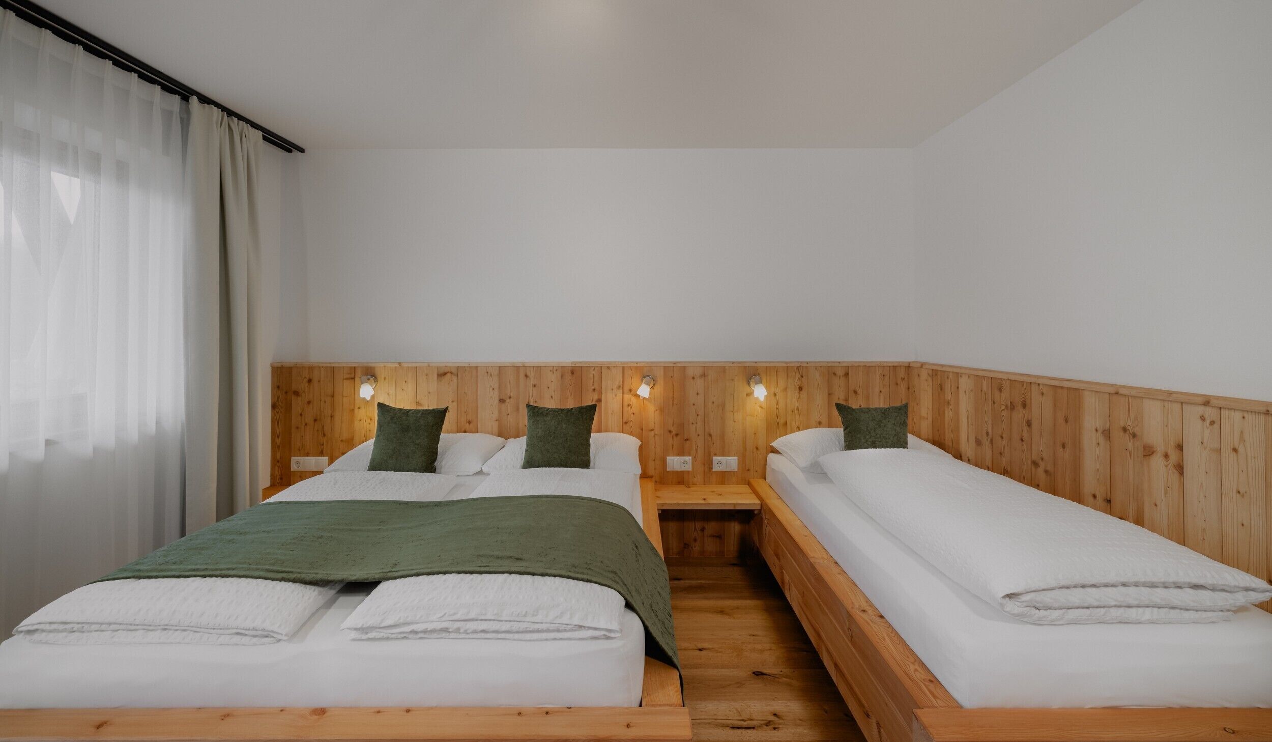 Une chambre avec deux lits, des têtes de lit en bois, du linge de lit blanc et une fenêtre ornée de rideaux légers.