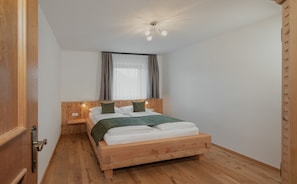 A wooden bed in a room with a wooden floor, a nightstand, a wall-mounted light, and a ceiling light fixture.