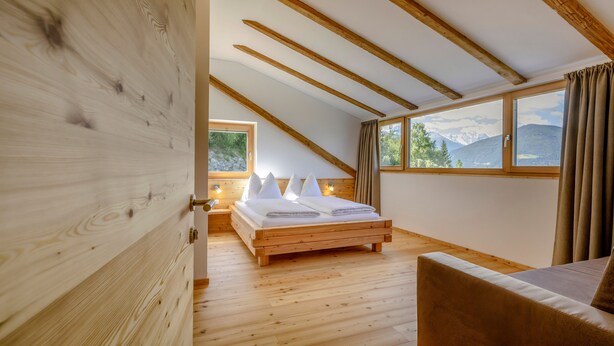 A bedroom with a wooden bed, a sofa, a window with a mountain view, and wooden beams on the ceiling. - Apartment 'Vernalhof Apt Panorama' with Mountain View, Shared Terrace and Wi-Fi (Sluderno)