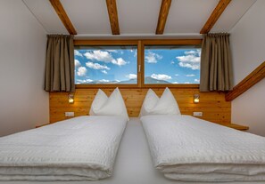 Une chambre avec deux lits, des poutres apparentes en bois au plafond, une grande fenêtre avec des rideaux et des lampes fixées au mur.