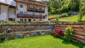 A multi-story building with balconies, a stone retaining wall, and a grassy area with flowers. - Apartment 'Vernalhof Apt Panorama' with Mountain View, Shared Terrace and Wi-Fi (Sluderno)
