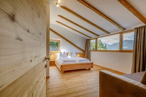 A bedroom with a wooden floor, a bed with white linens, a wooden ceiling, and a window with a mountain view.