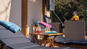 Una silla reclinable con cojín, una mesita de madera con un libro y dos frascos de mermelada, y una cesta de frutas al fondo.