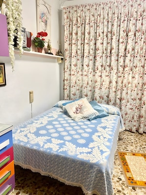 Une chambre à coucher comprenant un lit, une commode, une étagère avec des objets décoratifs et un rideau à motifs floraux.