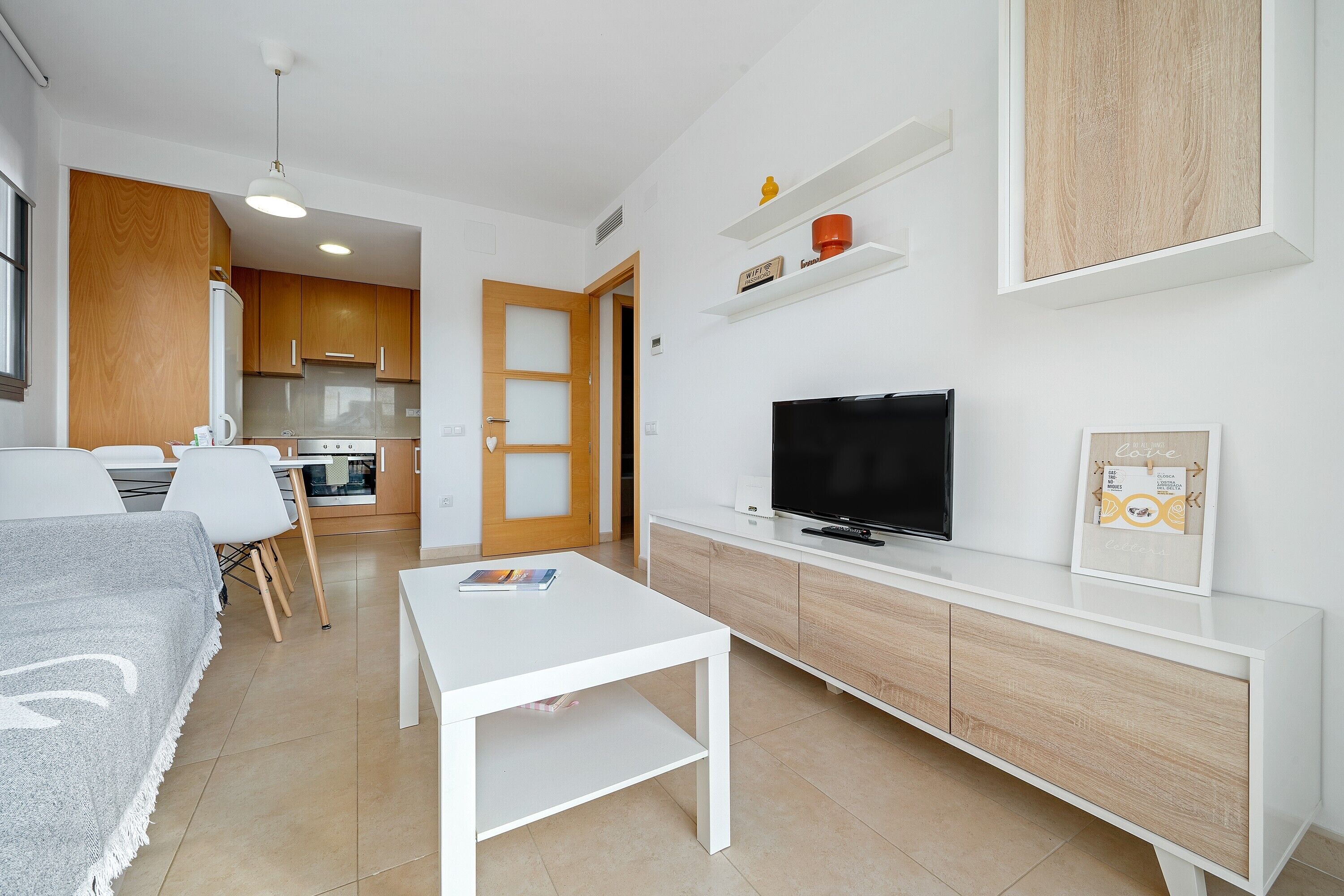 Una sala de estar moderna con un sofá blanco, una mesa de centro, un televisor y una cocina al fondo.