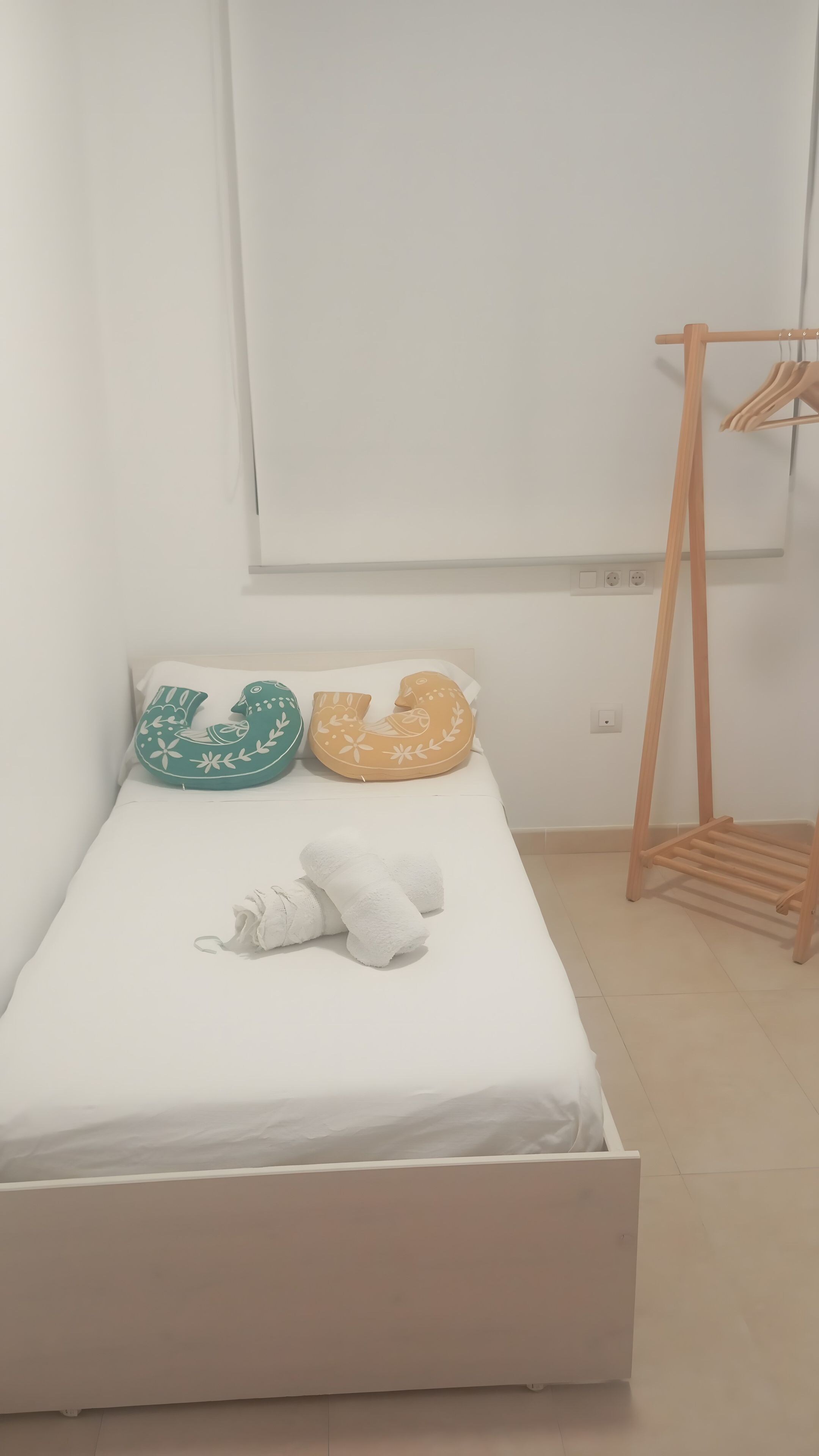 A single bed with a white mattress and two decorative pillows, a wooden hanger rack, and a plain white wall.