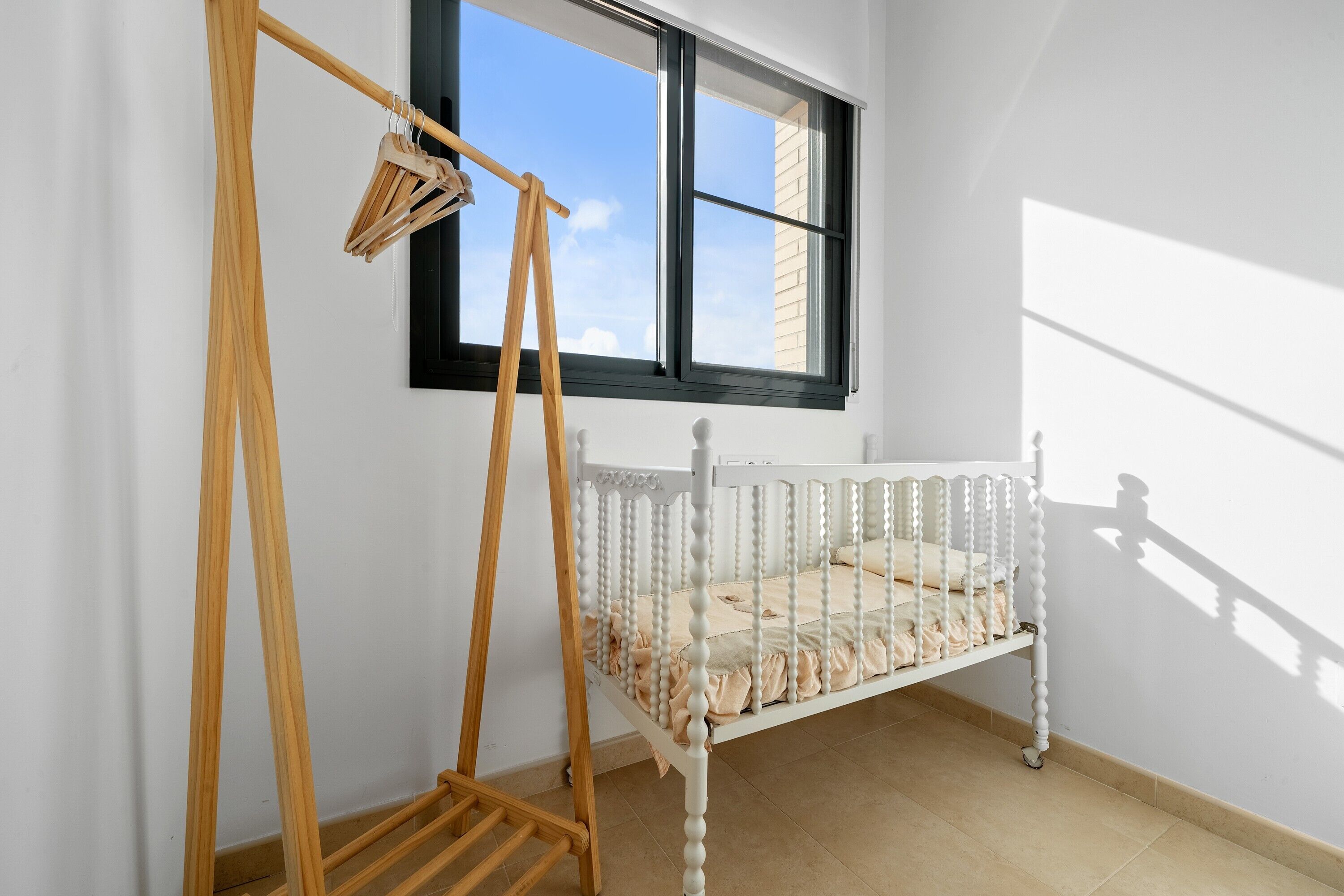 Een wit kinderbed met een gevlinderd matras, een houten schommel en een raam met zwarte kozijnen.