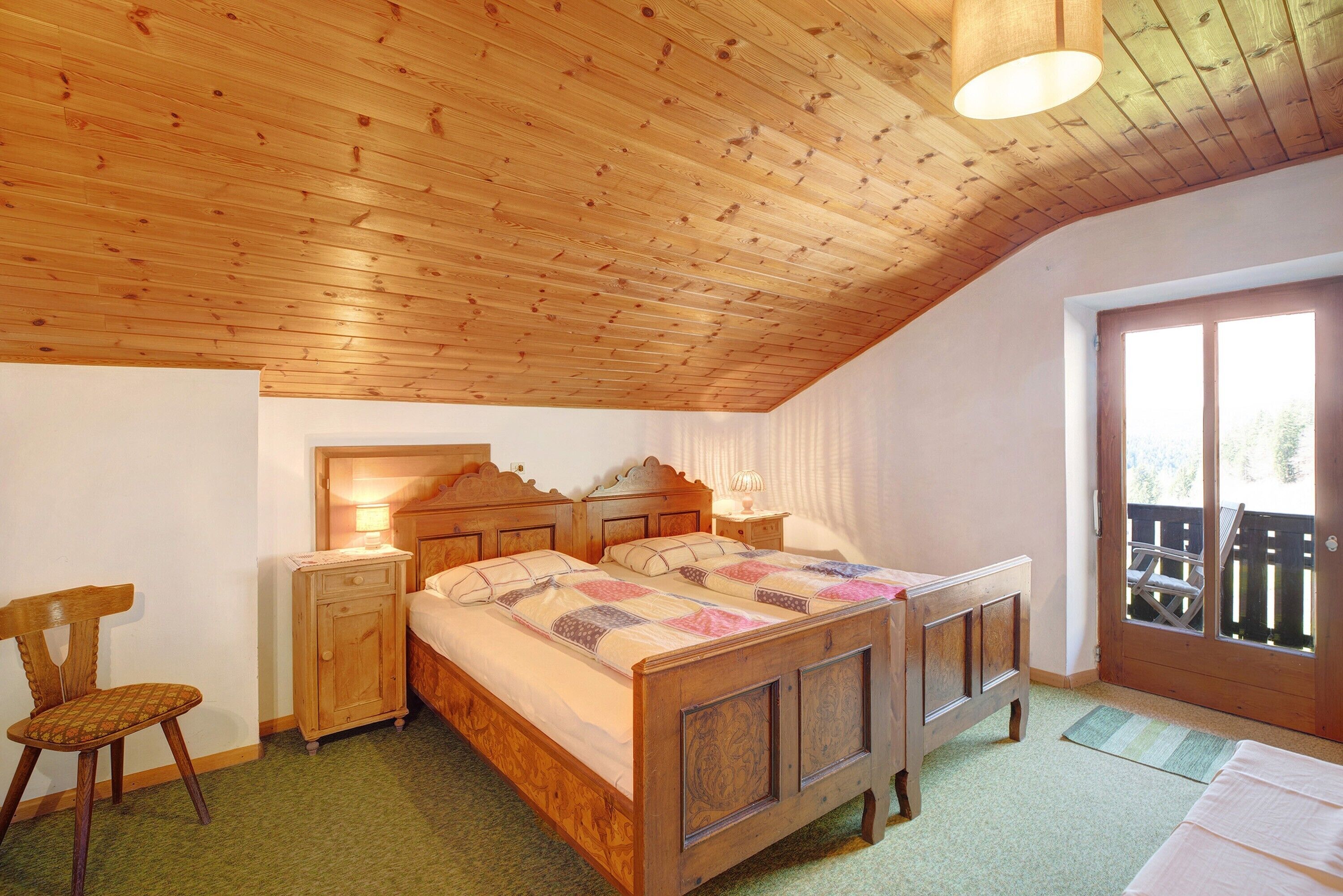 A bedroom with a wooden ceiling, a bed with a quilt, a nightstand, a chair, and a window with a view.