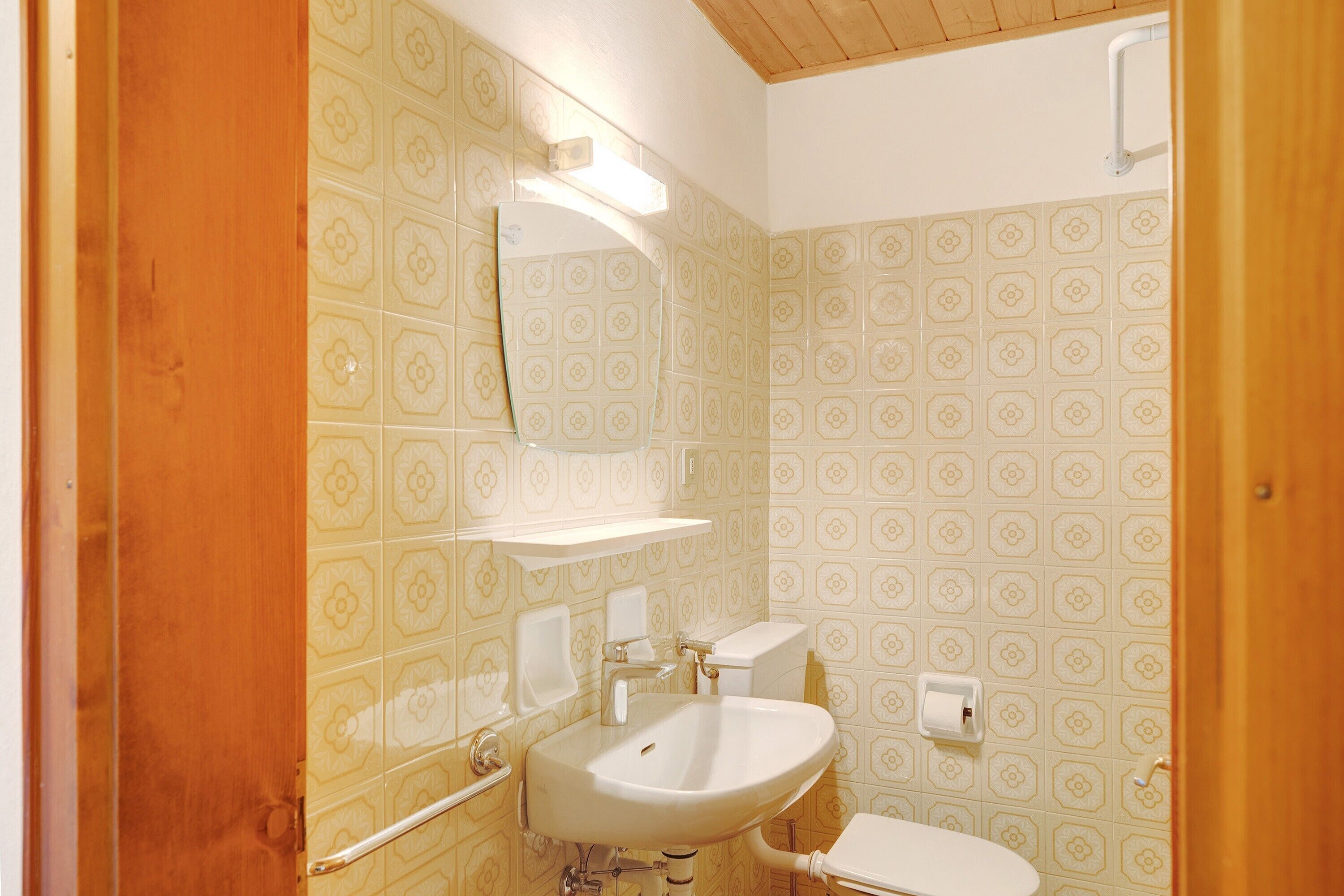 Casa de banho com lavatório, espelho, sanita e azulejos estampados.