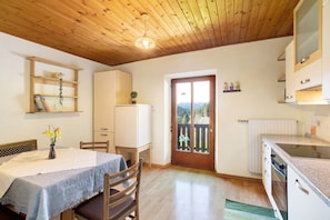 Une cuisine avec des meubles de rangement en bois, une table à manger avec des chaises, une étagère suspendue avec des livres et une vue à travers une porte.