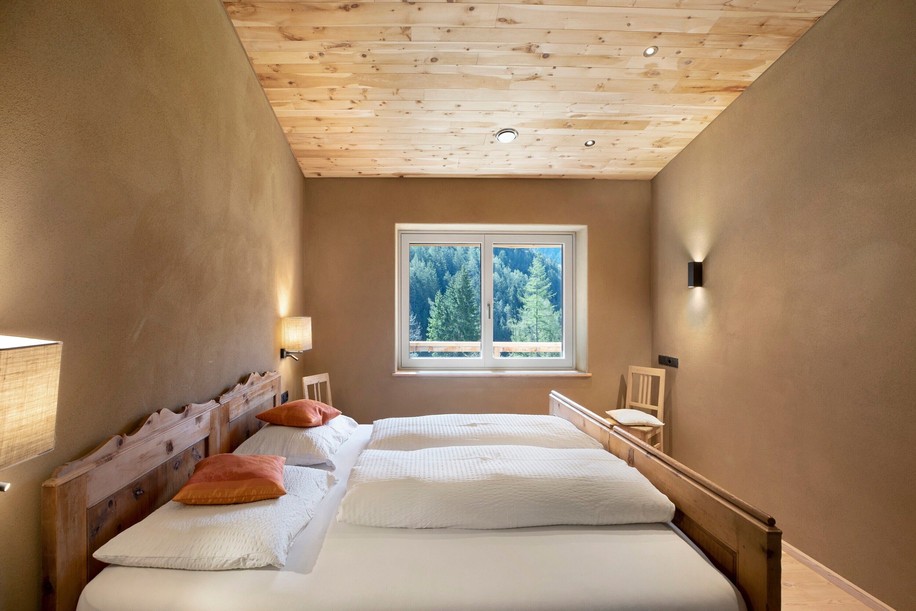 Une chambre avec un plafond en bois, une fenêtre donnant sur les arbres, un lit avec une literie blanche et une table de chevet avec une lampe.