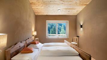 Une chambre avec un plafond en bois, une fenêtre donnant sur les arbres, un lit avec une literie blanche et une table de chevet avec une lampe.