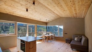 Een modern interieur met een houten plafond, grote ramen met uitzicht op een bos, een eethoek en een bank.