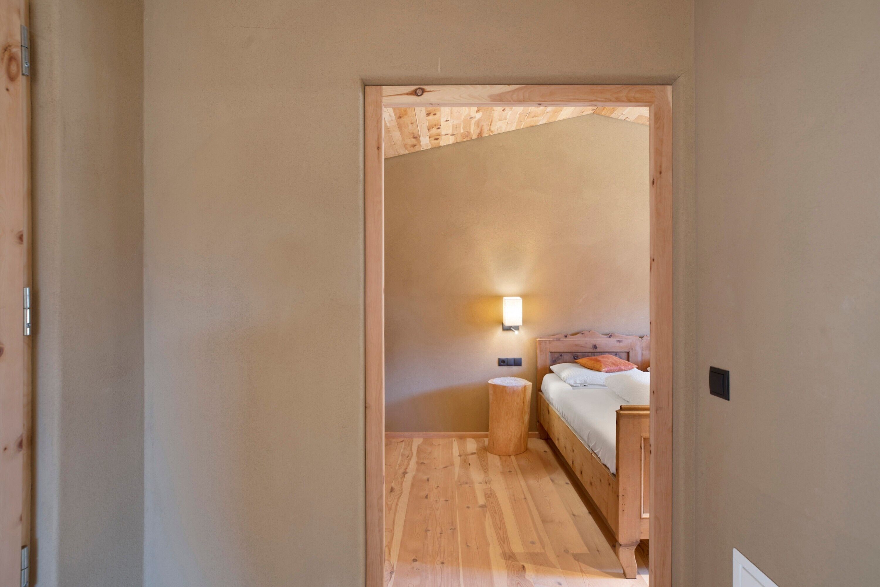 Un corridoio stretto che porta a una camera da letto con pavimento in legno, un letto con cuscini, un comodino e una lampada a muro.
