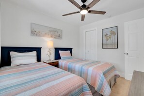 A bedroom with two beds, a ceiling fan, a nightstand with a lamp, and artwork on the walls.