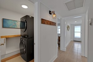 A laundry area with a washing machine and dryer, a wooden shelf with hooks, a wall-mounted light fixture, and a painting of boats on the wall.