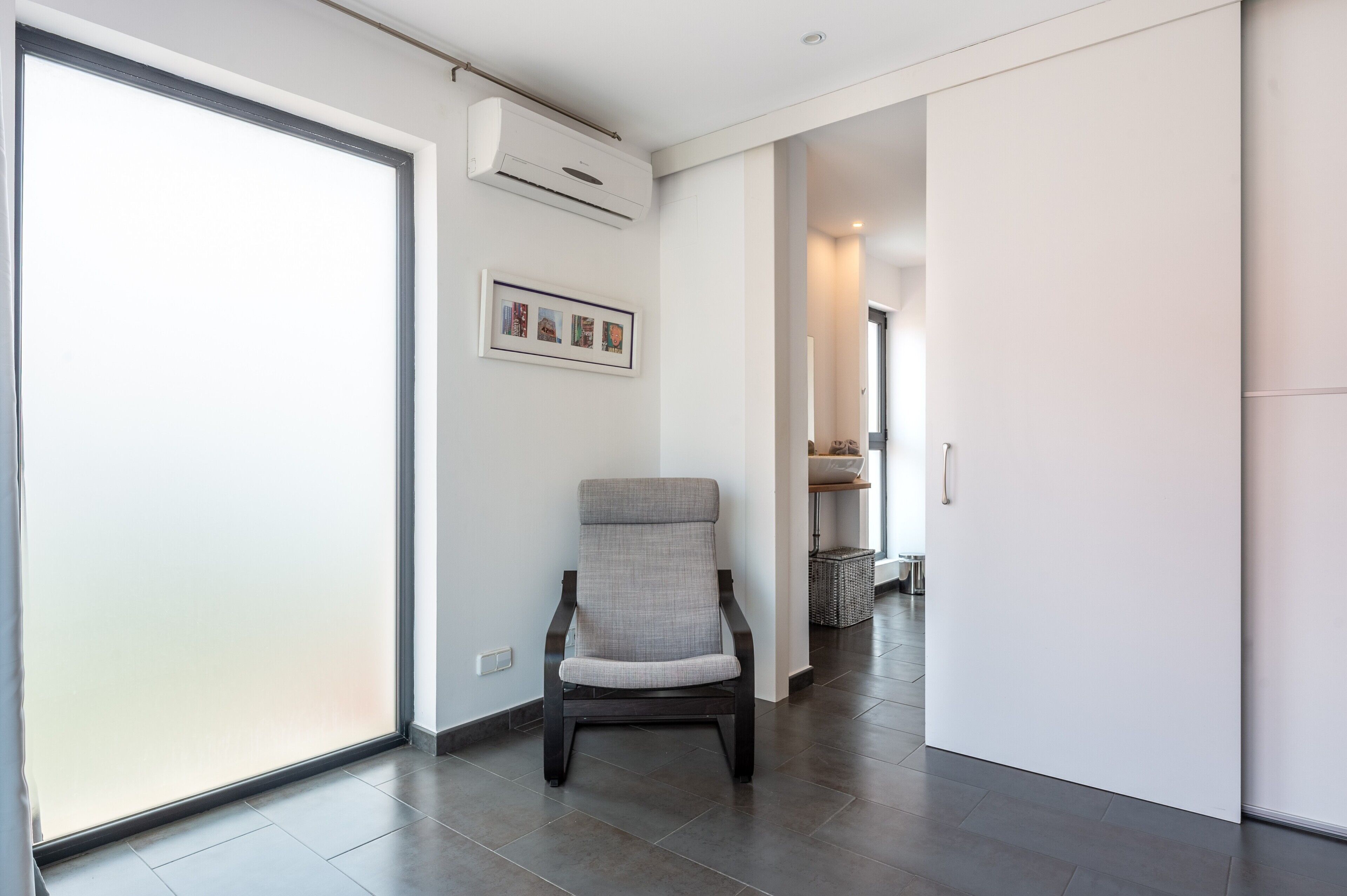 A modern interior with a sliding glass door, a grey upholstered chair, a framed picture on the wall, and a hallway leading to another room.