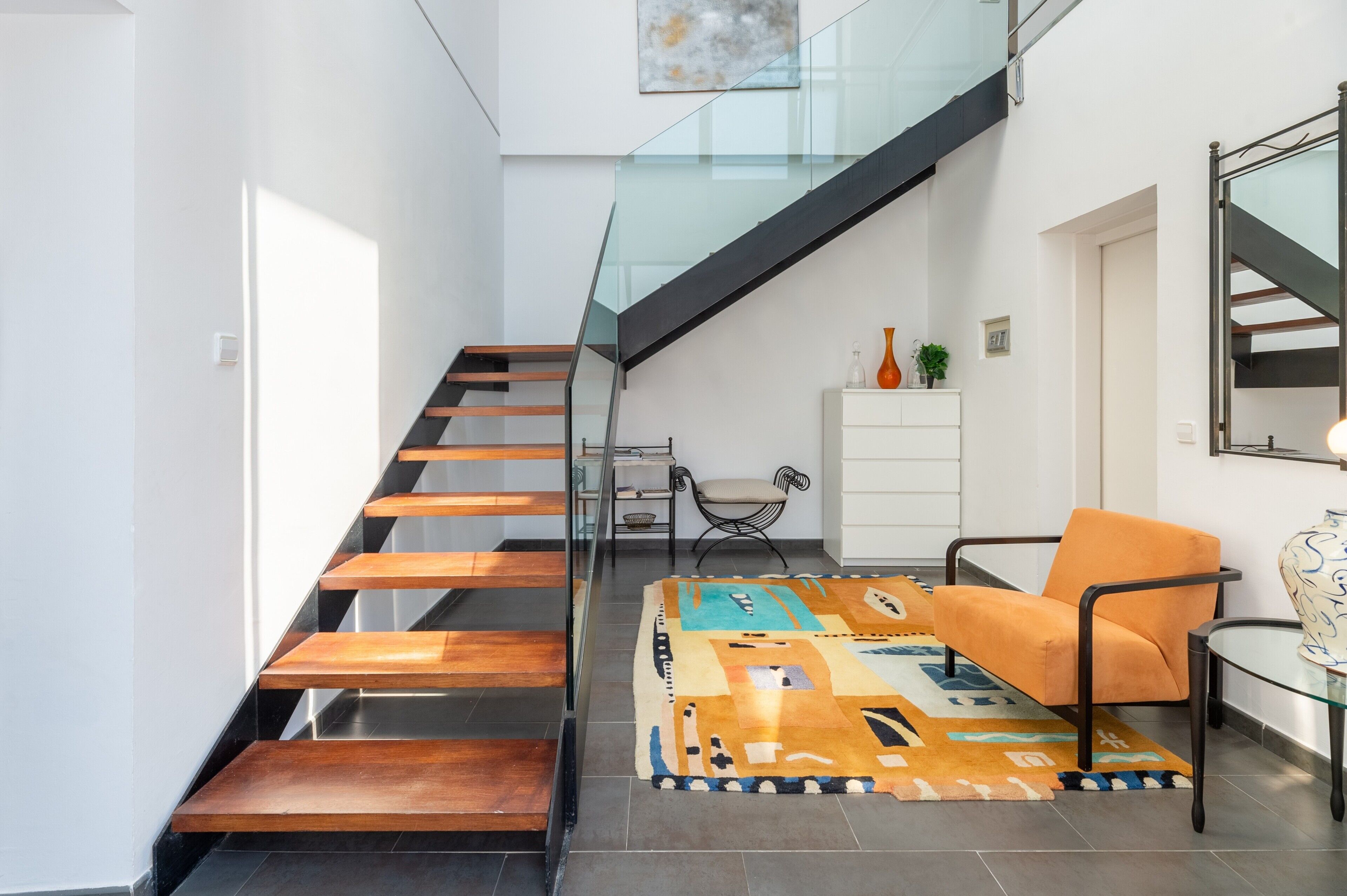 A modern interior with a glass staircase, a patterned rug, and a single orange chair.