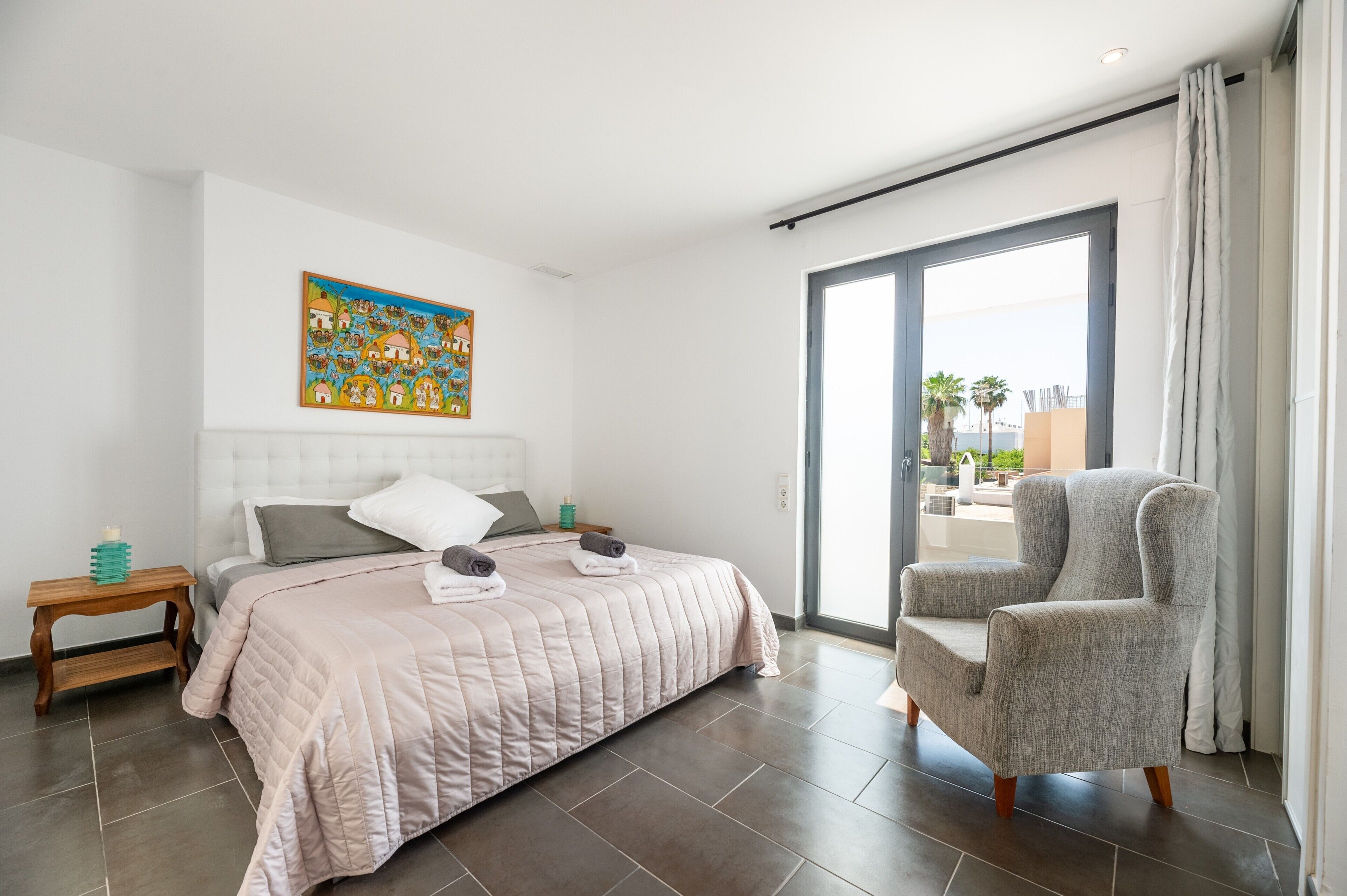 Ein Schlafzimmer mit Bett, Stuhl, Nachttisch, einem Gemälde und Blick durch eine Glastür.