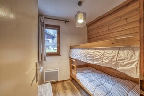 Une chambre avec un lit superposé doté d’une tête de lit en bois, une fenêtre avec des rideaux, un radiateur et un luminaire suspendu.