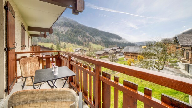 A balcony with a table and chairs, overlooking a village with houses and mountains.