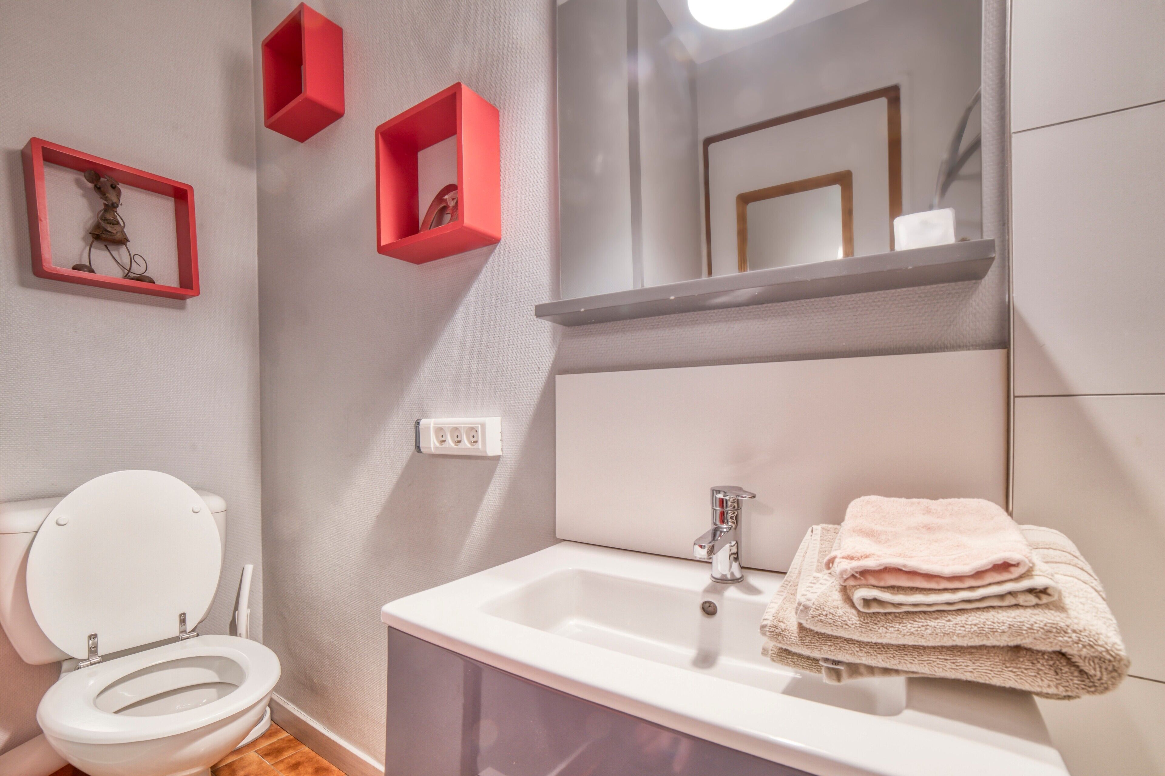 Une salle de bain avec des toilettes, un lavabo et un miroir. Il y a des boîtes de rangement rouges accrochées au mur.