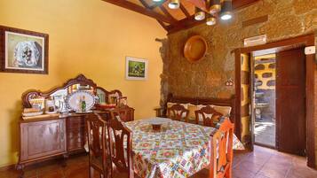 A dining area with a wooden table, chairs, and a patterned tablecloth. There is a wooden cabinet with a mirror and decorative items. The walls are yellow, and there is a stone wall with a wooden door.