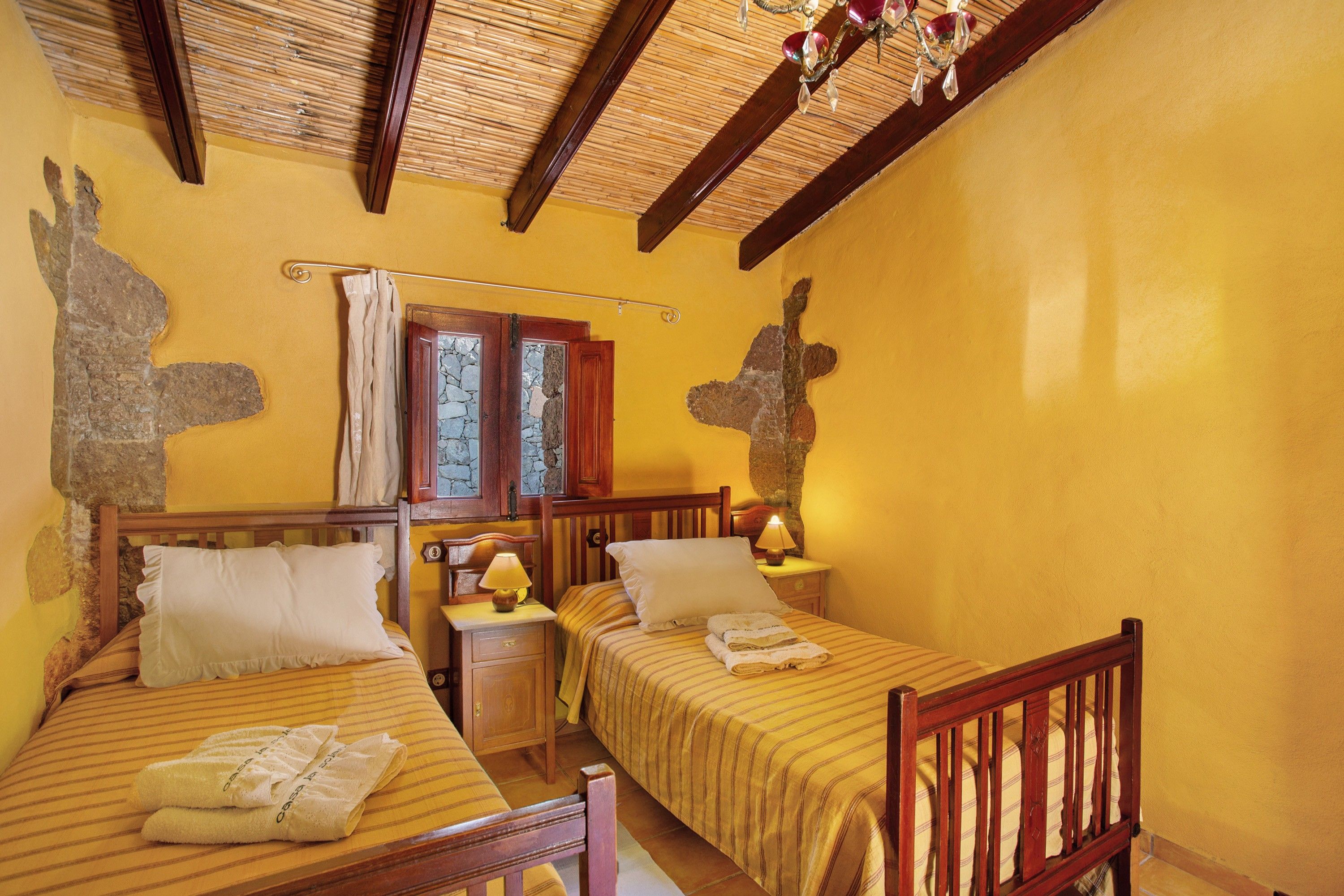A room with two beds, wooden headboards, a nightstand with a lamp, and a stone wall.