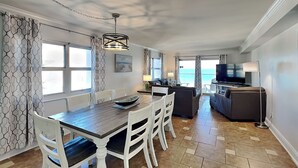 A modern room with a dining table, chairs, a sofa, a TV, and a balcony with a view of the sea. - Regency Towers #801 West | Beachfront, Pool! (Pensacola Beach)
