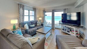 A living room with a sofa, a coffee table, and a TV. There is a balcony with a view of the ocean. - Regency Towers #801 West | Beachfront, Pool! (Pensacola Beach)