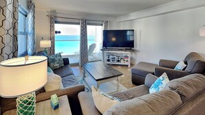 A modern living room with a large flat-screen TV, comfortable seating, and a view of the ocean. - Regency Towers #801 West | Beachfront, Pool! (Pensacola Beach)