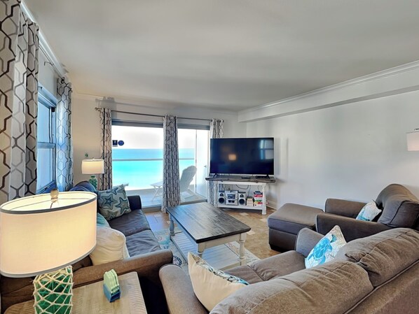 A modern living room with a large flat-screen TV, comfortable seating, and a view of the ocean.