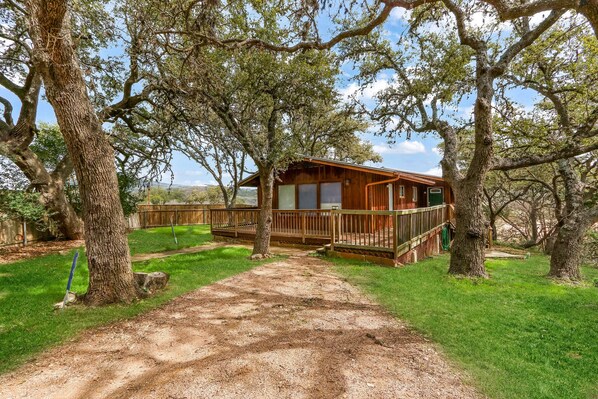A rustic cabin with a wooden deck, surrounded by large trees and a dirt path leading to the entrance.