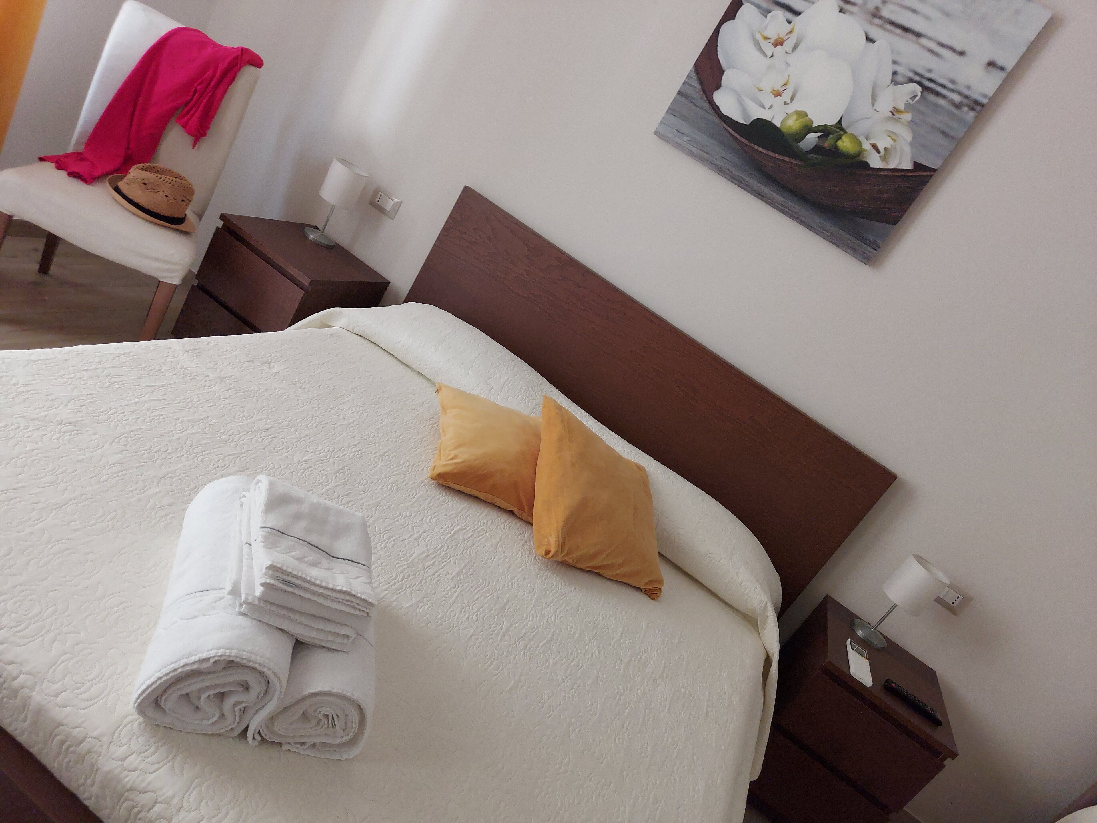 Una cama bien hecha con sábanas blancas y dos almohadas, una toalla doblada, una mesita de noche con una lámpara y un cuadro de orquídeas blancas colgado en la pared.
